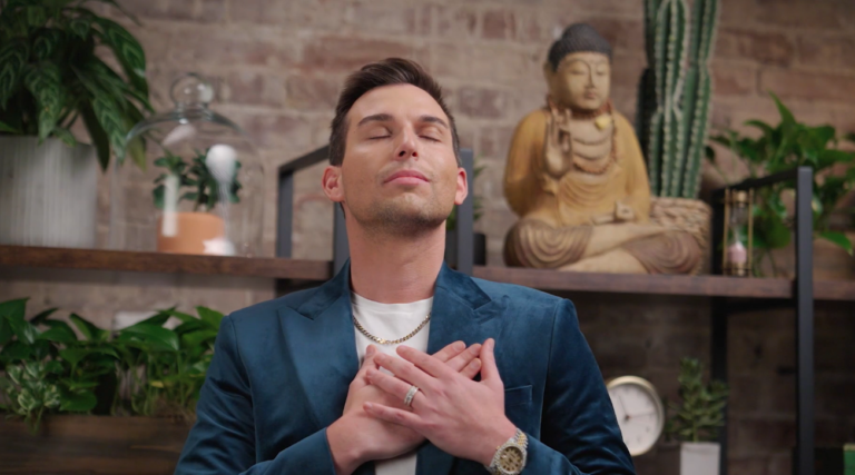 A person, Matt Fraser, in a blue blazer stands with closed eyes and hands over chest in a serene setting. A Buddha statue and plants are in the background.